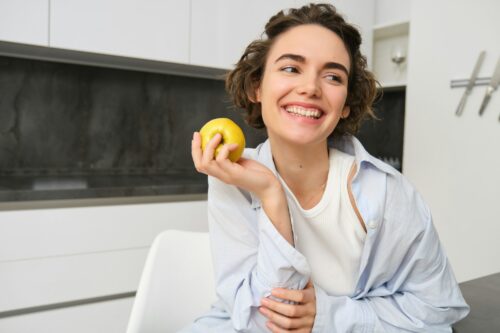 Image of brunette woman laughing, smiling while eating an apple in kitchen, sitting at home, having